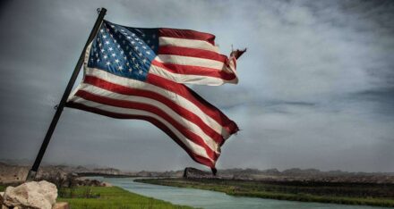 a damaged american flag flying on a pool and a river and clouds can be seen in background.