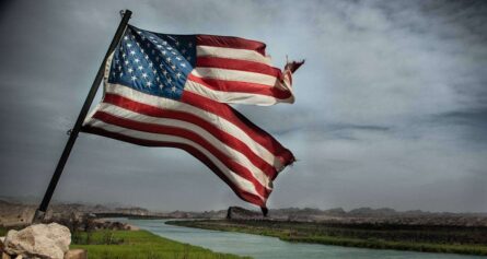 a damaged american flag flying on a pool and a river and clouds can be seen in background.